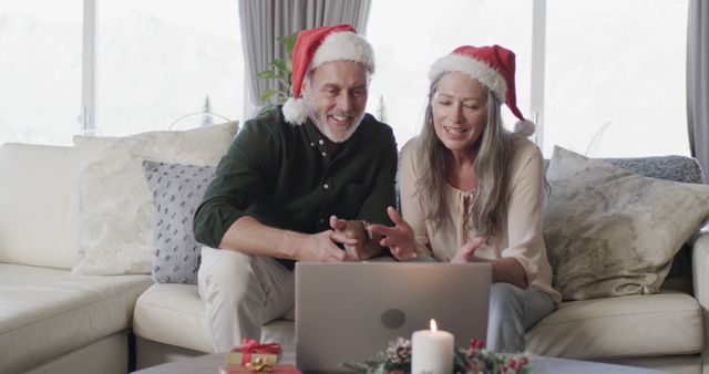 Senior couple wearing Santa hats is sitting on sofa making a video call on a laptop during Christmas holidays. Candles and gifts emphasize festive ambiance. Perfect for use in stories about holiday celebrations, technology in communication, and cozy home environments.
