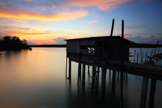 Rustic lakeside shack on stilts captured at sunset, reflecting in calm waters. Ideal for depicting tranquility, remote living, and peaceful natural landscapes. Perfect for promotional use in travel, relaxation, photography contexts, and nature-related articles.