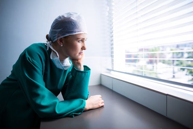 Female surgeon in surgical attire standing by a window, looking outside with a thoughtful expression. Ideal for use in articles about healthcare professionals, medical field challenges, mental health of medical staff, or hospital environments.