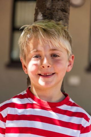 Smiling blonde boy in red-and-white striped shirt standing by tree trunk, sunny portrait