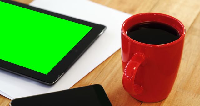 Digital tablet featuring green screen on wooden desk next to red coffee cup. Ideal for concepts related to technology use, remote work environments, office setup, breakfast routines, and productivity strategies.