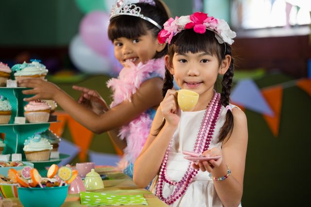 Two young girls are enjoying a tea party during a birthday celebration. They are dressed up with accessories like tiaras, feather boas, and beaded necklaces. The table is decorated with colorful cupcakes and party decorations. This image is perfect for use in content related to children's parties, celebrations, and playful activities.