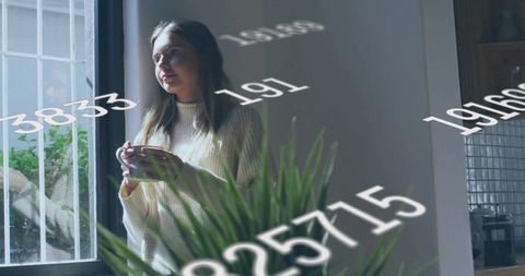 Holding mug and gazing through barred window with floating numbers overlay in cozy kitchen