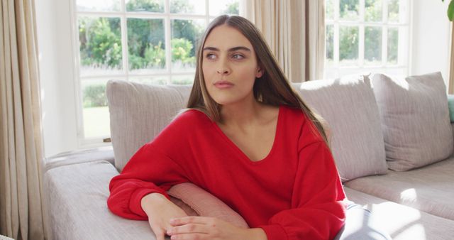 Young woman sitting on couch in living room, wearing red sweater. Large window in background brings in natural light, creating calm and relaxing atmosphere. Great for topics on relaxation, lifestyle, home decor, and leisure activities.