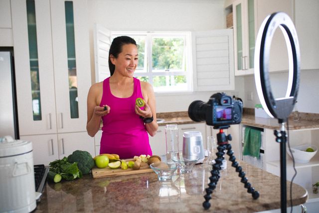 Asian female vlogger smiling while preparing healthy juice with fresh fruits in a modern kitchen. Ideal for content related to healthy living, cooking tutorials, influencer marketing, and home-based vlogging setups. Useful for promoting kitchen appliances, healthy recipes, and social media engagement.