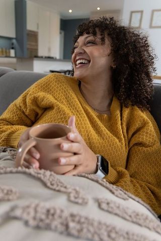 Woman in 30s relaxing on sofa with mug and blanket in cozy open-plan kitchen