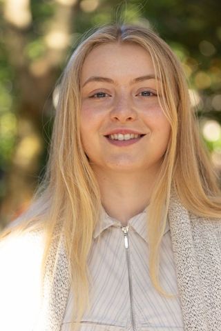 Smiling young woman outdoors in sunlit park wearing striped zip shirt and knit cardigan