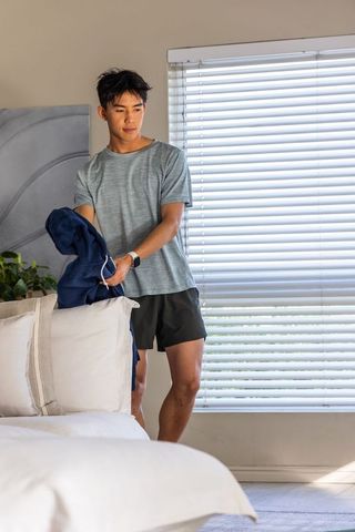 Asian man checking smartwatch while holding navy jacket in minimalist bedroom morning light