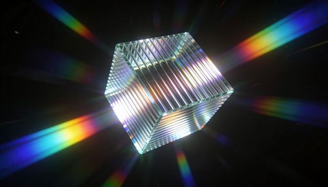 Prismatic ridged glass cube scattering vivid rainbow light beams across dark background