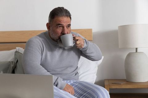 Mature man sipping coffee on bed while using laptop, morning routine in modern bedroom