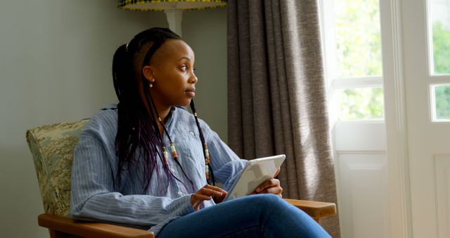 Side view of young black woman using digital tablet and sitting on the couch in comfortable home. She is looking away 4k