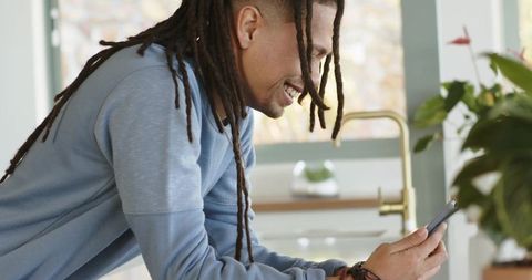 Smiling man with dreadlocks leaning on kitchen counter using smartphone in modern home