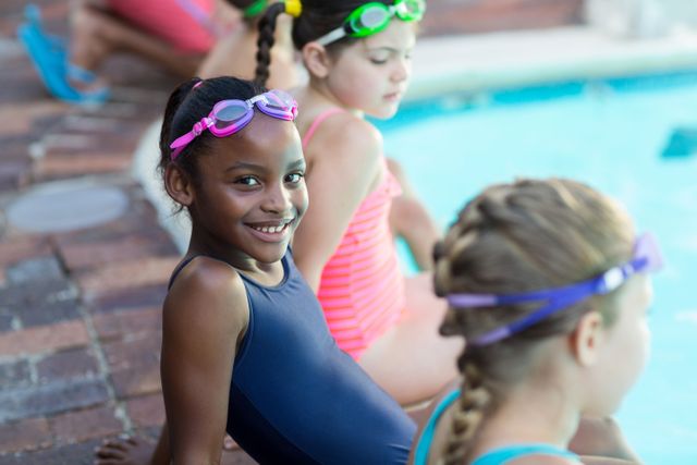 Young girl smiling while sitting with friends by the pool. They are wearing swim goggles and swimwear, enjoying a summer day. Ideal for use in advertisements for summer camps, swimming lessons, children's activities, and outdoor fun.