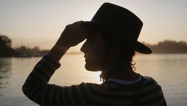 Backlit silhouette woman holding fedora at lakeshore during golden hour sunset
