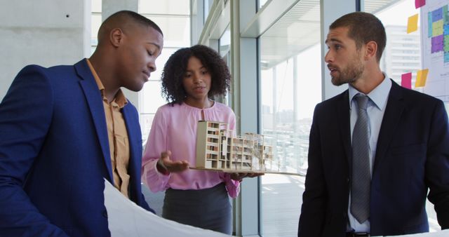 This image shows an architect team engaged in the discussion of a building model and blueprints in a modern office. This image can be used for websites, presentations, and marketing materials related to architecture, urban development, business collaboration, and professional teamwork. It depicts diversity, modern workplace dynamics, and innovative practices.