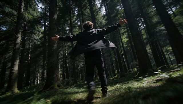 Man striding into sunlit conifer forest with outstretched arms, motion blur, moody woods