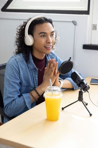 Non-binary creator speaking into microphone wearing white headphones in bright home studio