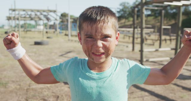 Boy with dirt on face and shirt showing muscles, standing in outdoor obstacle course. Ideal for content on children's sports, physical fitness, determination, summer activities, and overcoming challenges. Suitable for use in school programs, youth sports promotions, and motivational materials.
