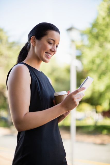 Businesswoman standing outdoors, smiling while using smartphone. Holding coffee cup, dressed in professional attire. Ideal for business, communication, technology, and lifestyle themes.