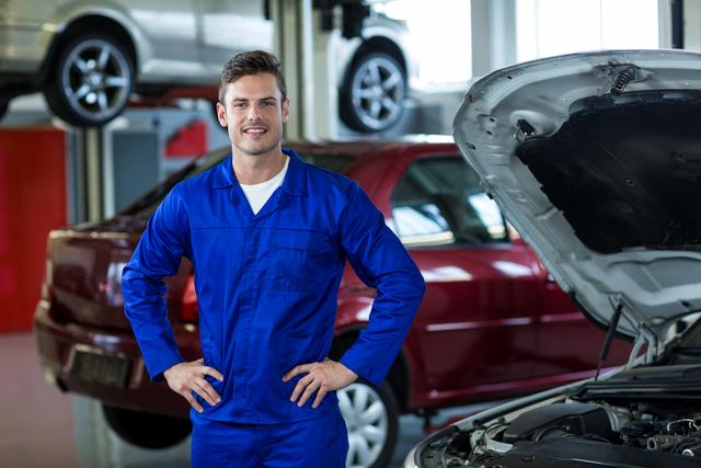 Mechanic in blue overalls standing confidently with hands on hips in an auto repair shop. Ideal for use in advertisements for automotive services, repair shops, and professional mechanic services. Can also be used in articles or blogs about car maintenance, automotive careers, and professional services.