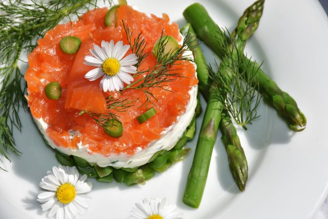 Smoked salmon rests on a layer of cream cheese, garnished with fresh asparagus spears and adorned with small flowers and dill. Suitable for depicting gourmet cuisine, restaurant menus, catering services, culinary blogs, or cooking class promotions.