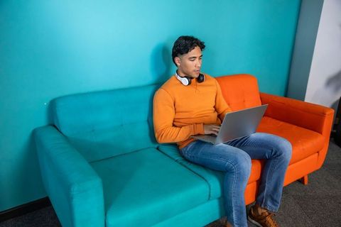 Young man working remotely on laptop on vibrant teal and orange sofa wearing headphones