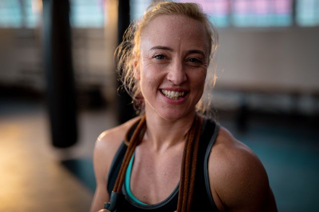 This image captures a smiling Caucasian woman in a gym setting during her workout. She is wearing sports clothes and appears to be in the middle of a strength training or boxing session. This image can be used for promoting fitness programs, gym memberships, healthy lifestyle campaigns, or motivational content related to exercise and well-being.