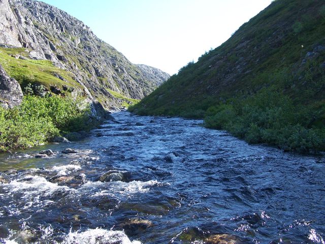 Mountain stream flowing through a rocky valley under bright daylight. Clear water is surrounded by lush greenery and rugged rocks with hills in background. Great for illustrating outdoor activities, nature exploration, and scenic beauty. Ideal for travel brochures, outdoor adventure promotions, and environmental awareness campaigns.