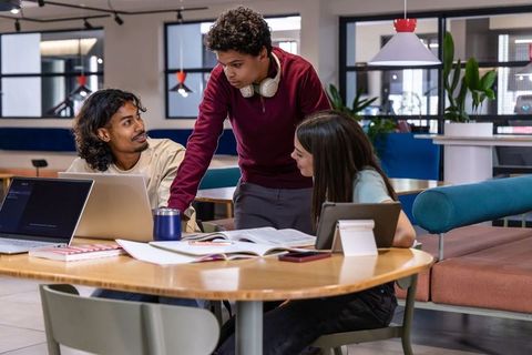 Students collaborating around round wooden table in modern coworking lounge with laptops