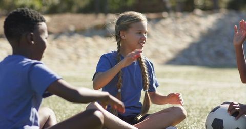 Sunlit multiracial youth sitting on grass passing soccer ball, practicing teamwork