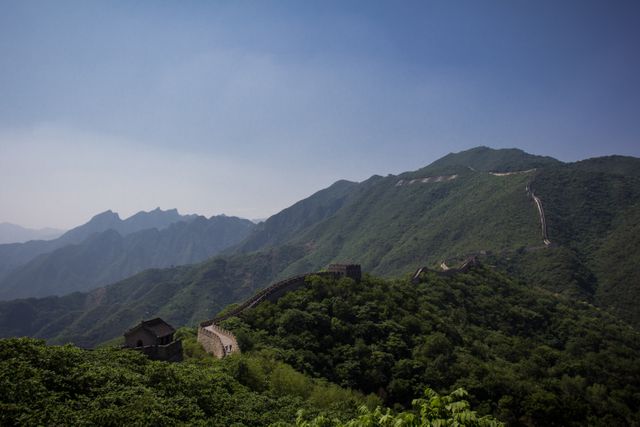 Great Wall of China extending through lush green mountains under a clear blue sky. Ideal for use in travel articles, tourism guides, cultural heritage publications, history documentaries, and nature-focused content showcasing stunning scenic views and landmark sites.