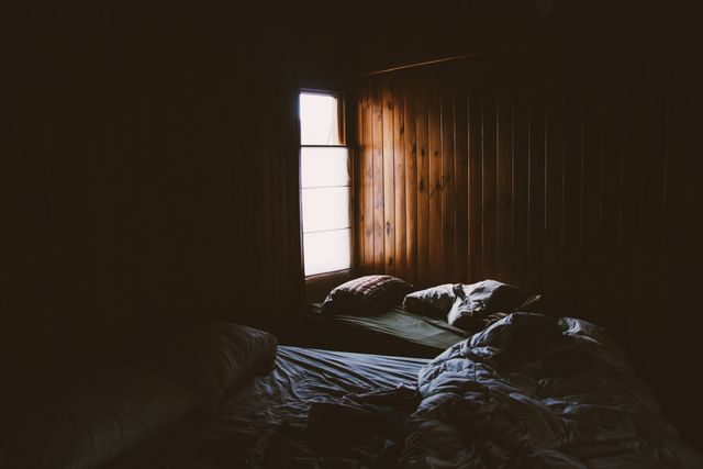 Depicts a rustic bedroom with light streaming through a window onto wooden walls and unmade beds. Ideal for use in designs emphasizing cozy cabins, tranquility, or relaxed mornings. Eye-catching for nature retreats, interior design showcasing rustic style or promoting stress-free living environments.