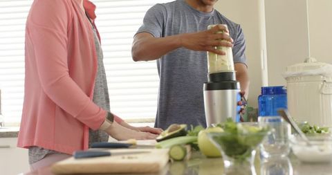 Couple preparing fresh green smoothie with personal blender on bright kitchen island