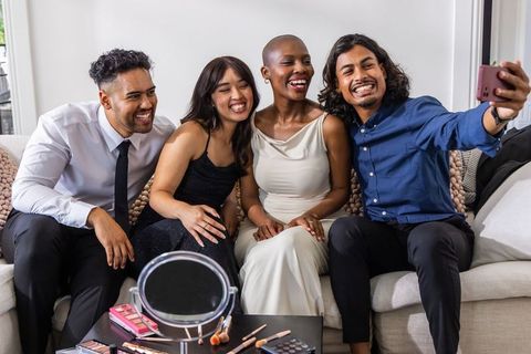 Diverse friends dressed up smiling and taking selfie on sofa with makeup and smartphone