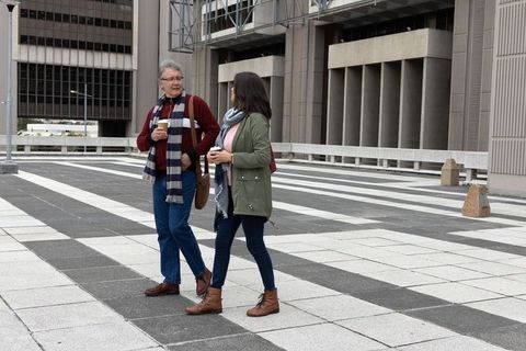 Senior couple walking across urban plaza holding coffee cups, chatting near concrete building