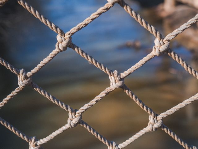 Close-up view of a knotted rope net against a blurred water background. Ideal for use in themes related to safety, protection, nautical activities, and outdoor environments. The texture and pattern can serve as an interesting visual element for backgrounds, designs, or advertisements focused on securing spaces.
