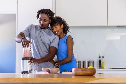 African american couple preparing fresh fruit smoothie in modern bright kitchen together