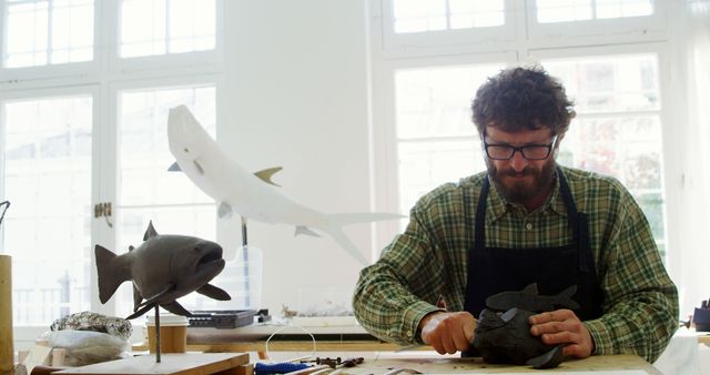 Man wearing glasses and checkered shirt, working meticulously on a fish sculpture in a brightly lit workshop. Surroundings include small tools and wooden models of fish, suggesting a focus on artisanal craftsmanship. Ideal for themes of creativity, hobbies, woodworking, and handmade craftsmanship.