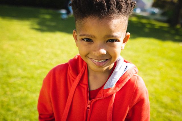 This image captures a joyful African American boy smiling at the camera while standing outdoors in a backyard. The boy is wearing a red hoodie, and the bright green grass and sunny day add to the cheerful atmosphere. This image is perfect for use in advertisements, educational materials, family-oriented content, and articles about childhood, happiness, and outdoor activities.