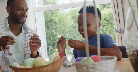 African american father and son painting colorful easter eggs at sunlit kitchen table