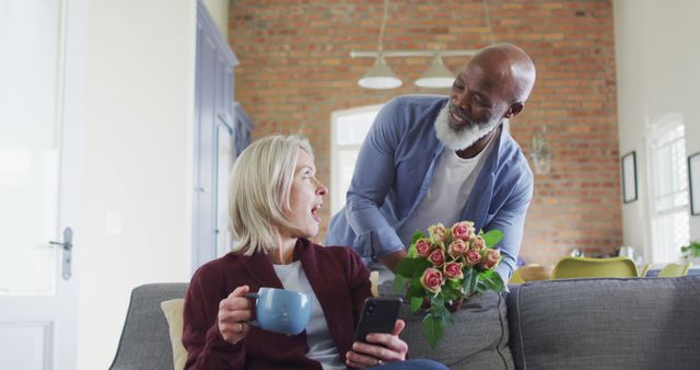 Elderly man surprising his partner with a bouquet of roses while she holds a mug and a smartphone, both in a cozy living room setting. Great for use in themes related to love, relationships, companionship, home lifestyle, and special occasions.