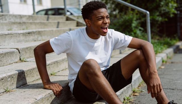 Laughing male teen sitting on steps in white tee and black shorts, outdoor urban lifestyle