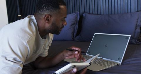 Midlife man working on laptop in bedroom writing in notebook and planning day