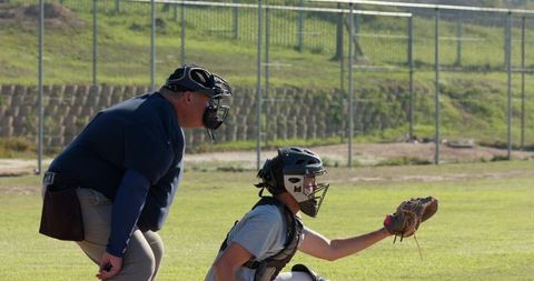 Baseball catcher crouching with mitt while umpire watching behind plate on sunny field with chainlin