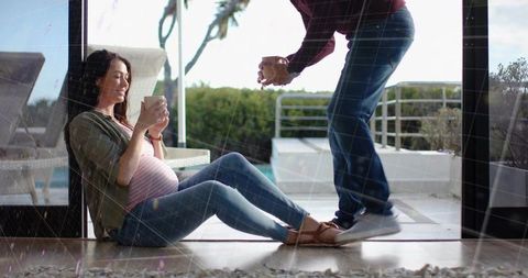 Expectant woman cradling mug and relaxing by patio door while partner offering drink