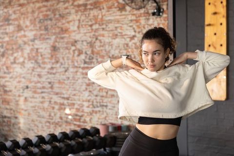 Asian woman lifting cropped top before workout in industrial gym with dumbbell rack