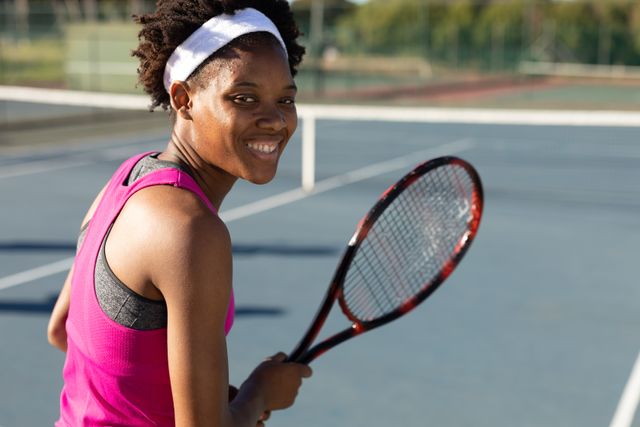 Young African American woman playing tennis on a sunny day, smiling and holding a tennis racket. Suitable for use in content promoting sports, competitions, fitness, active lifestyles, and athletic apparel.