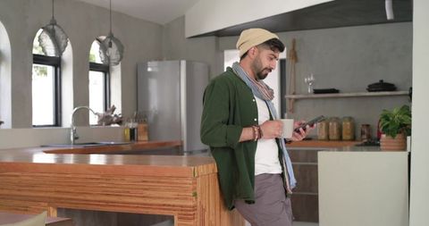 Indian man leaning on wood kitchen island holding smartphone and mug in modern rustic kitchen