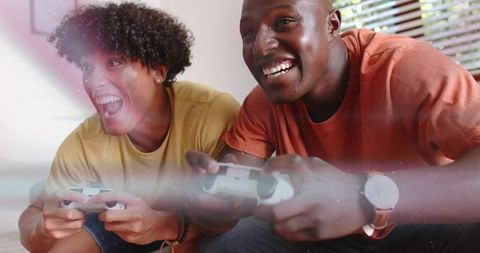 Two men playing video games on sofa laughing and competing with controllers in sunlit room