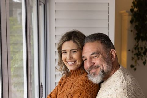 Mature Couple Embracing Beside Window Wearing Knit Sweaters, Cozy Home Togetherness